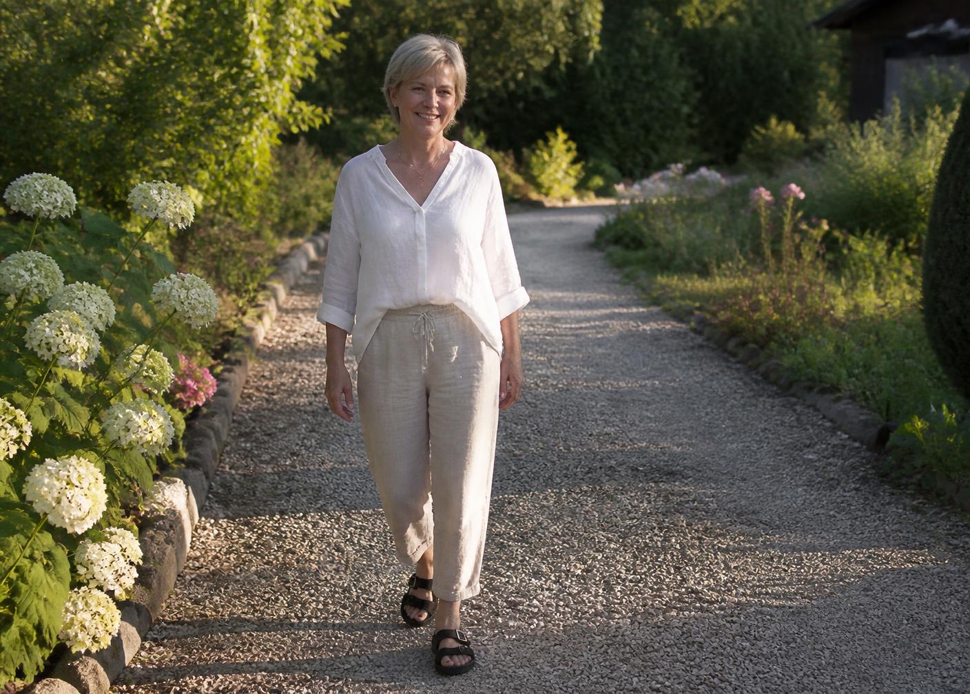 Frau um die 58 Jahre spaziert in Leinenkleidung und ergonomischen Sandalen über einen Gartenweg
